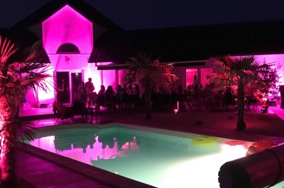 Piscine éclairée de nuit avec des lumières rouges et violettes, entourée de palmiers et d'une table avec un verre.