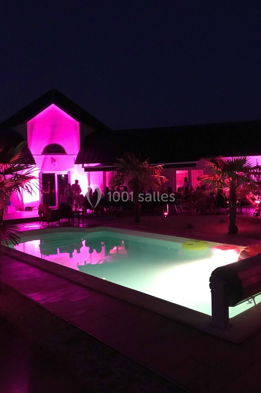 Piscine éclairée entourée de palmiers et d'une maison illuminée en rose, de nuit.