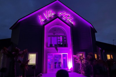 Piscine éclairée de nuit avec des lumières rouges et violettes, entourée de palmiers et d'une table avec un verre.