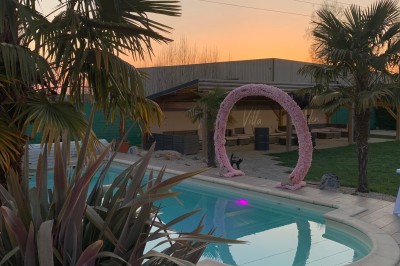 Piscine éclairée de nuit avec des lumières rouges et violettes, entourée de palmiers et d'une table avec un verre.