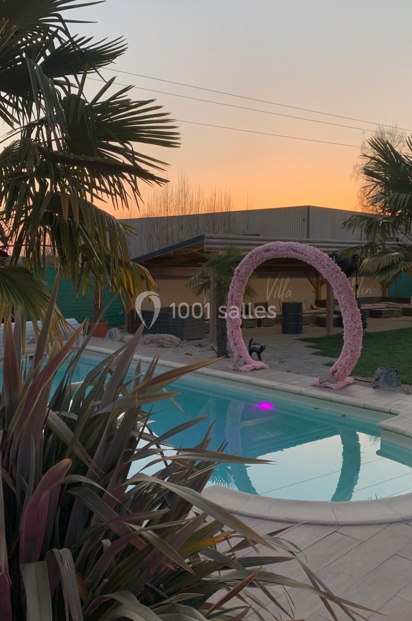 Piscine extérieure entourée de palmiers, avec une arche décorative et un coucher de soleil en arrière-plan.