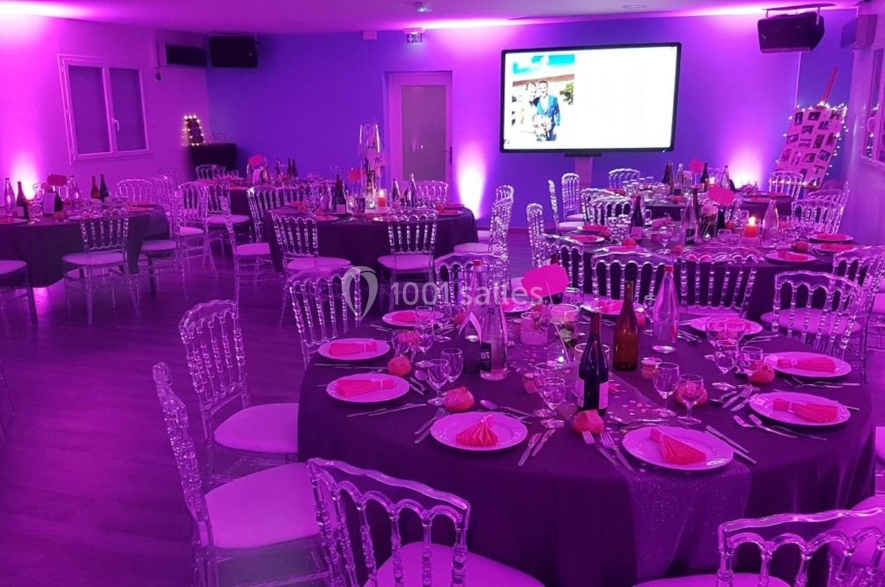 Salle de réception décorée avec des tables rondes dressées, éclairage violet et écran mural allumé.