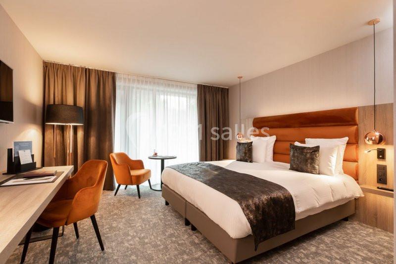 Chambre d'hôtel moderne avec lit double, bureau, fauteuils orange, grande fenêtre et éclairage chaleureux.