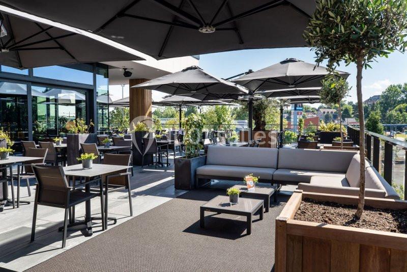 Terrasse extérieure aménagée avec tables, chaises, canapés et parasols, entourée de plantes et baignée de lumière naturelle.
