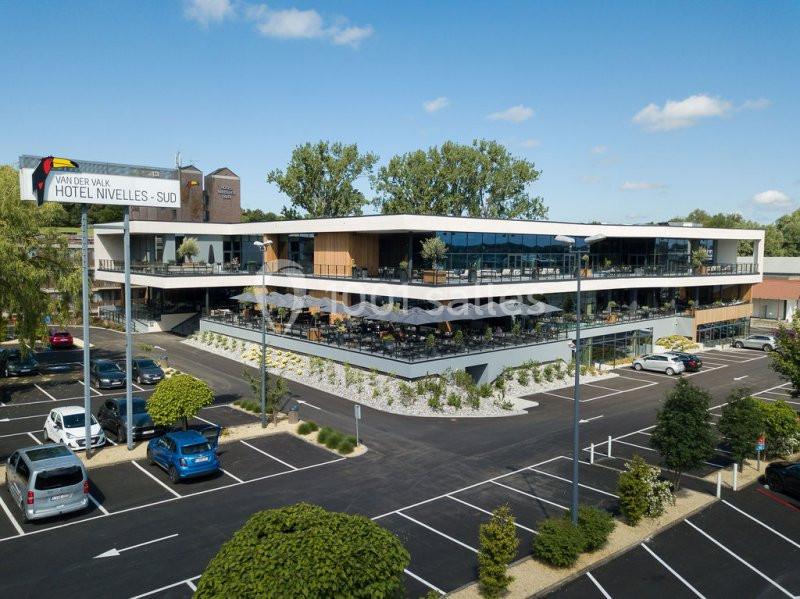 Vue extérieure d'un hôtel moderne à deux étages avec terrasses, entouré d'un parking et de végétation.