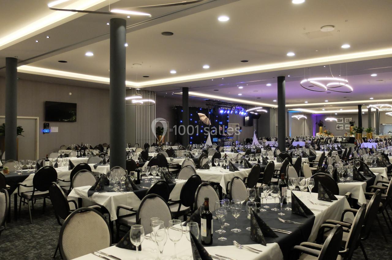 Salle de réception élégante avec tables dressées, nappes blanches, serviettes noires et scène éclairée en arrière-plan.