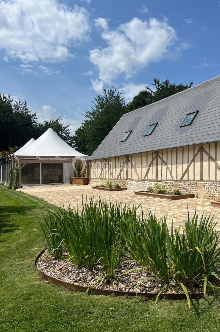 Cour pavée avec une maison à colombages, un jardin fleuri et une tente blanche sous un ciel partiellement nuageux.