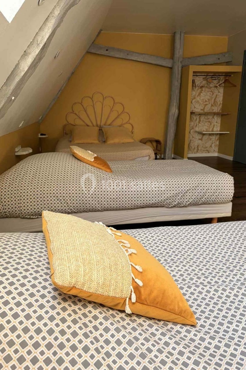 Chambre mansardée avec deux lits, murs jaunes, poutres apparentes et coussins assortis sur une literie à motifs.