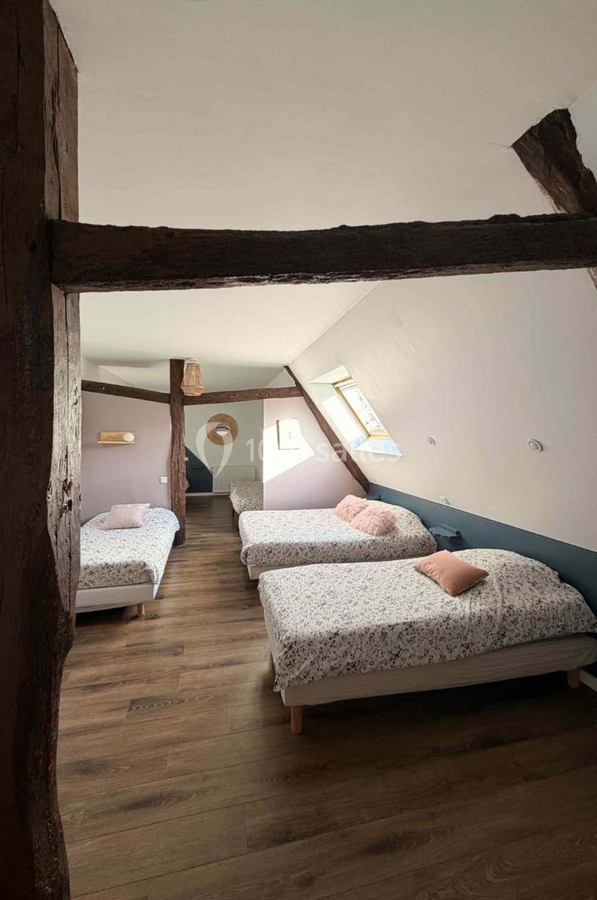 Chambre mansardée avec trois lits simples, poutres apparentes, parquet en bois et lumière naturelle.