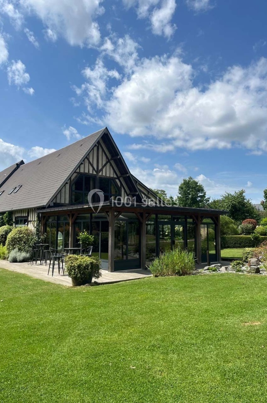 Maison à colombages avec véranda moderne, entourée de pelouse, arbustes et ciel bleu parsemé de nuages.
