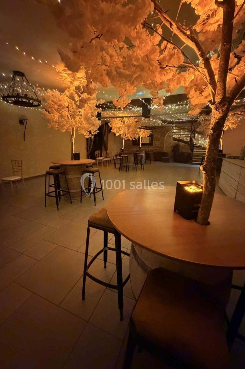 Salle décorée avec des arbres lumineux, des tables hautes et des guirlandes suspendues créant une ambiance chaleureuse.