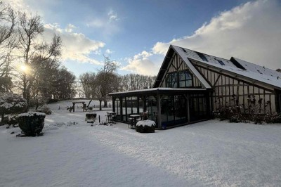 Des personnes discutent debout autour de tables en bois devant des tentes blanches dans un espace extérieur arboré.