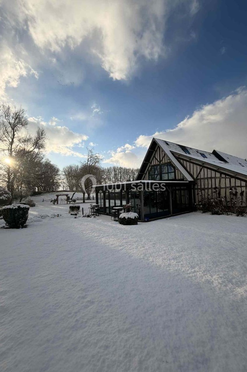 Maison à colombages entourée d'un jardin enneigé sous un ciel partiellement nuageux avec un soleil bas.