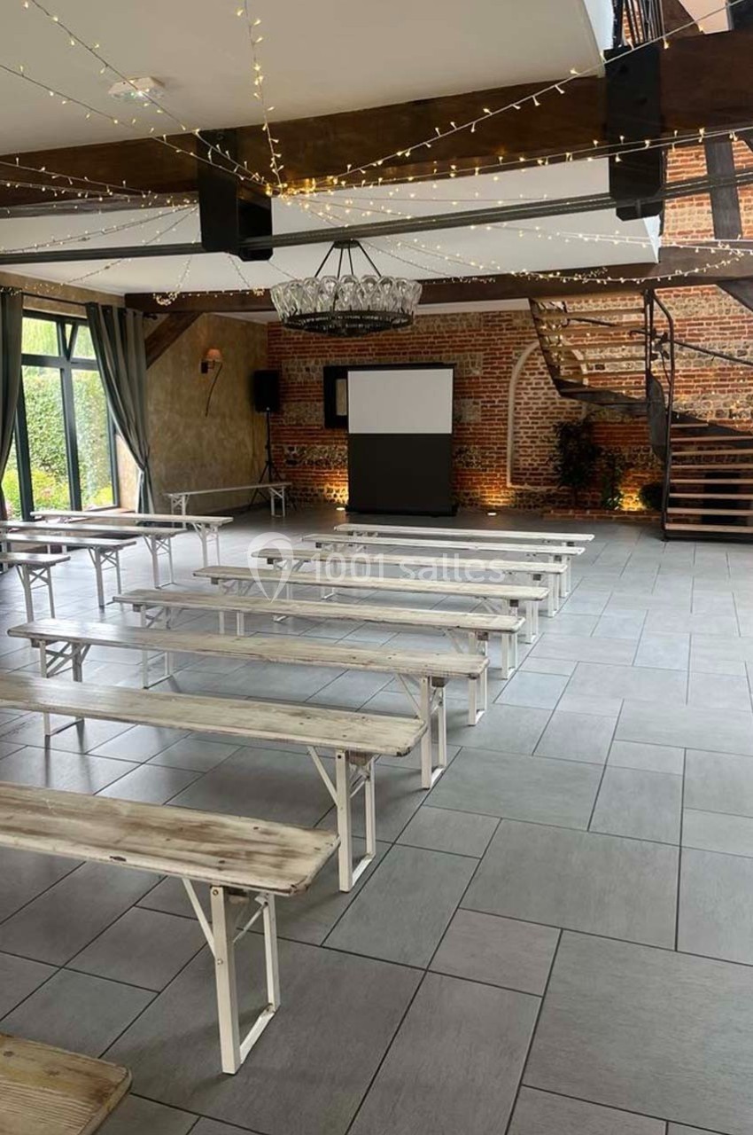 Salle lumineuse avec bancs en bois alignés, écran de projection, escalier en bois et murs en briques apparentes.
