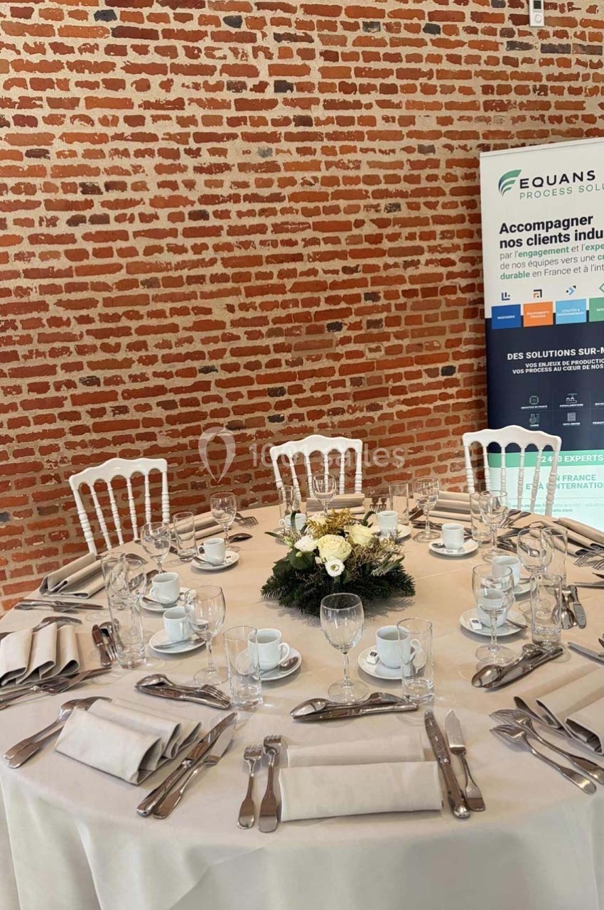 Table ronde dressée avec nappes blanches, vaisselle, couverts et centre de table floral, devant un mur en briques.