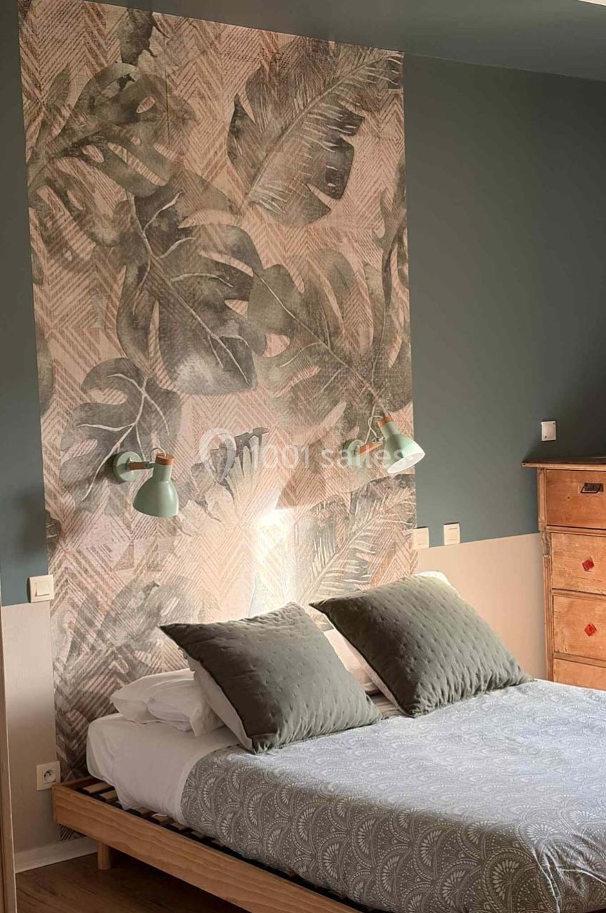 Lit double avec coussins gris, tête de lit décorée de motifs végétaux, éclairée par deux appliques murales vertes.