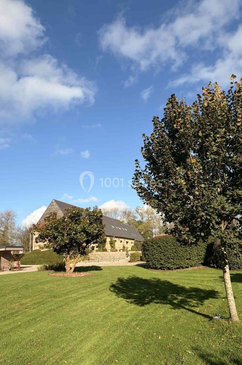 Pelouse verdoyante avec un arbre au premier plan, une maison en pierre et un ciel bleu parsemé de nuages.