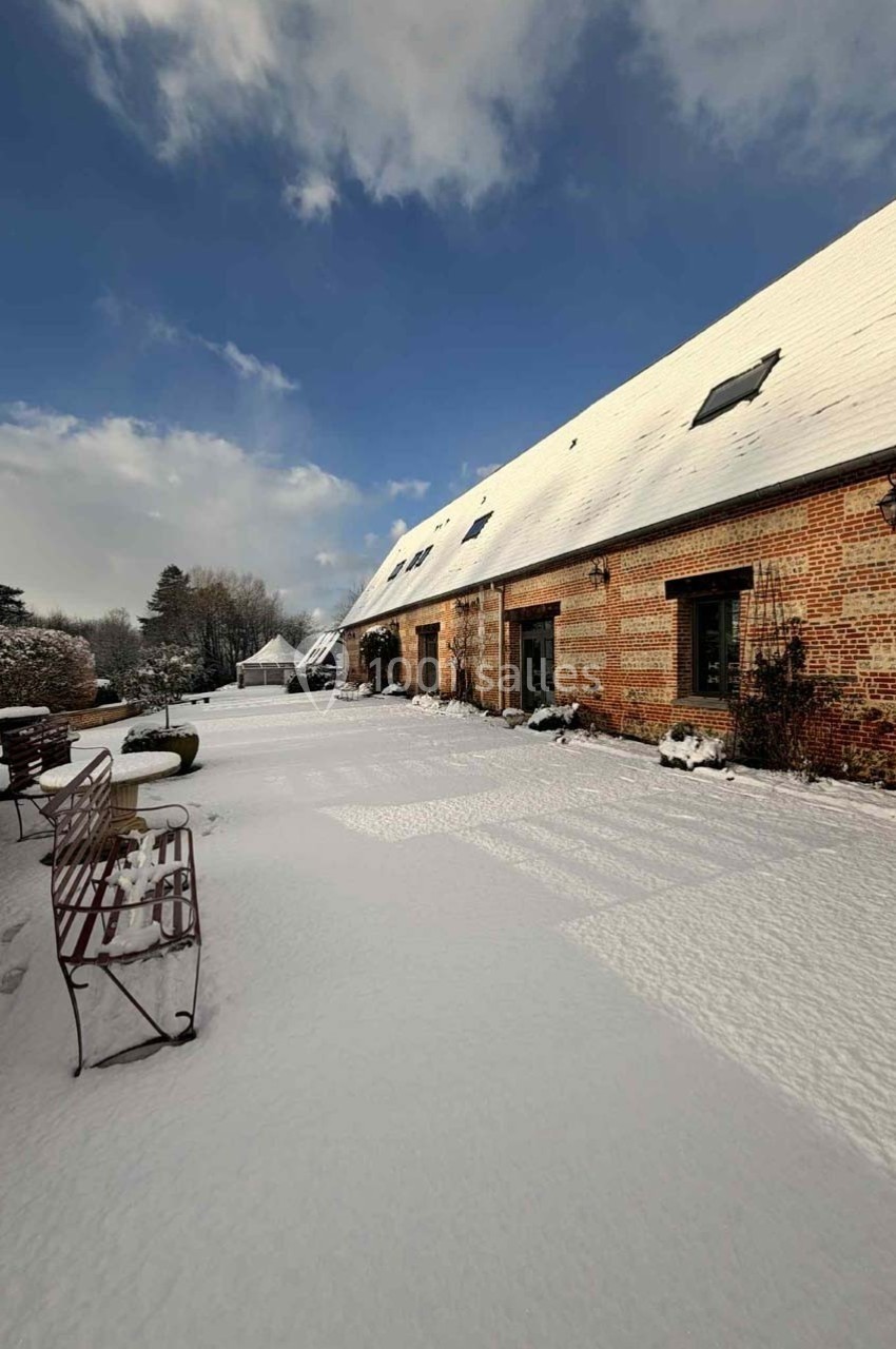 Cour d'une maison en briques sous la neige, avec bancs métalliques et ciel partiellement nuageux.
