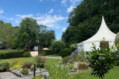 Des personnes discutent debout autour de tables en bois devant des tentes blanches dans un espace extérieur arboré.