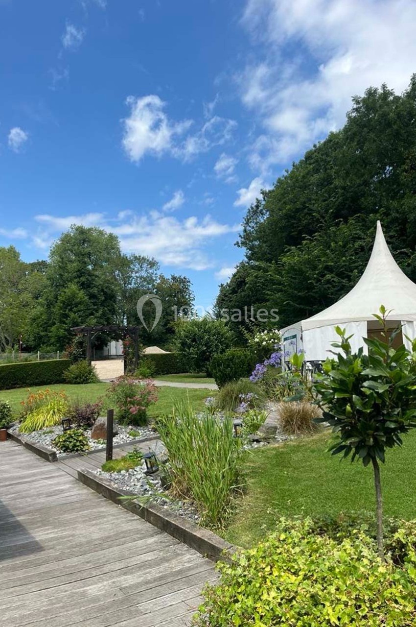 Jardin paysager avec pelouse, parterre fleuri, allée en bois et tente blanche sous un ciel bleu partiellement nuageux.