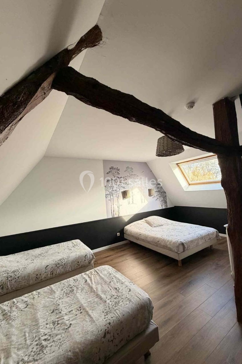 Chambre mansardée avec trois lits, poutres apparentes, parquet et fenêtre de toit laissant entrer la lumière naturelle.