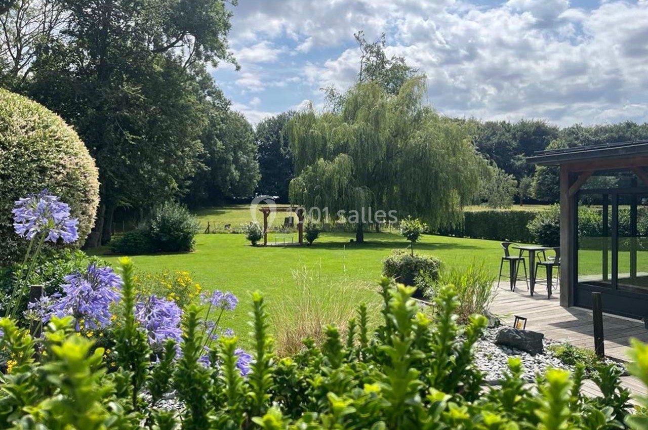 Jardin verdoyant avec pelouse, arbres, fleurs violettes au premier plan et une terrasse en bois avec mobilier.