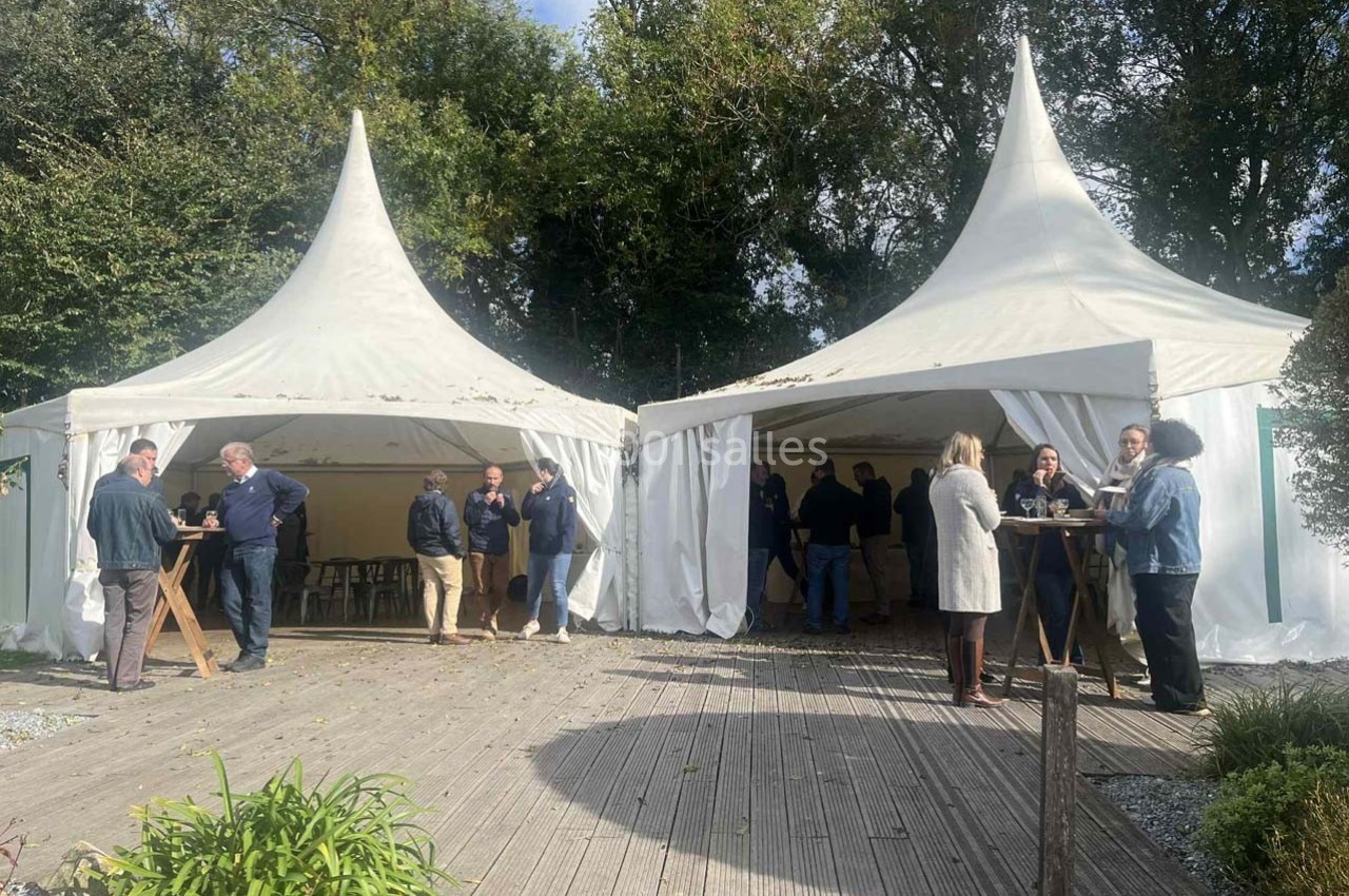 Des personnes discutent debout autour de tables en bois devant des tentes blanches dans un espace extérieur arboré.
