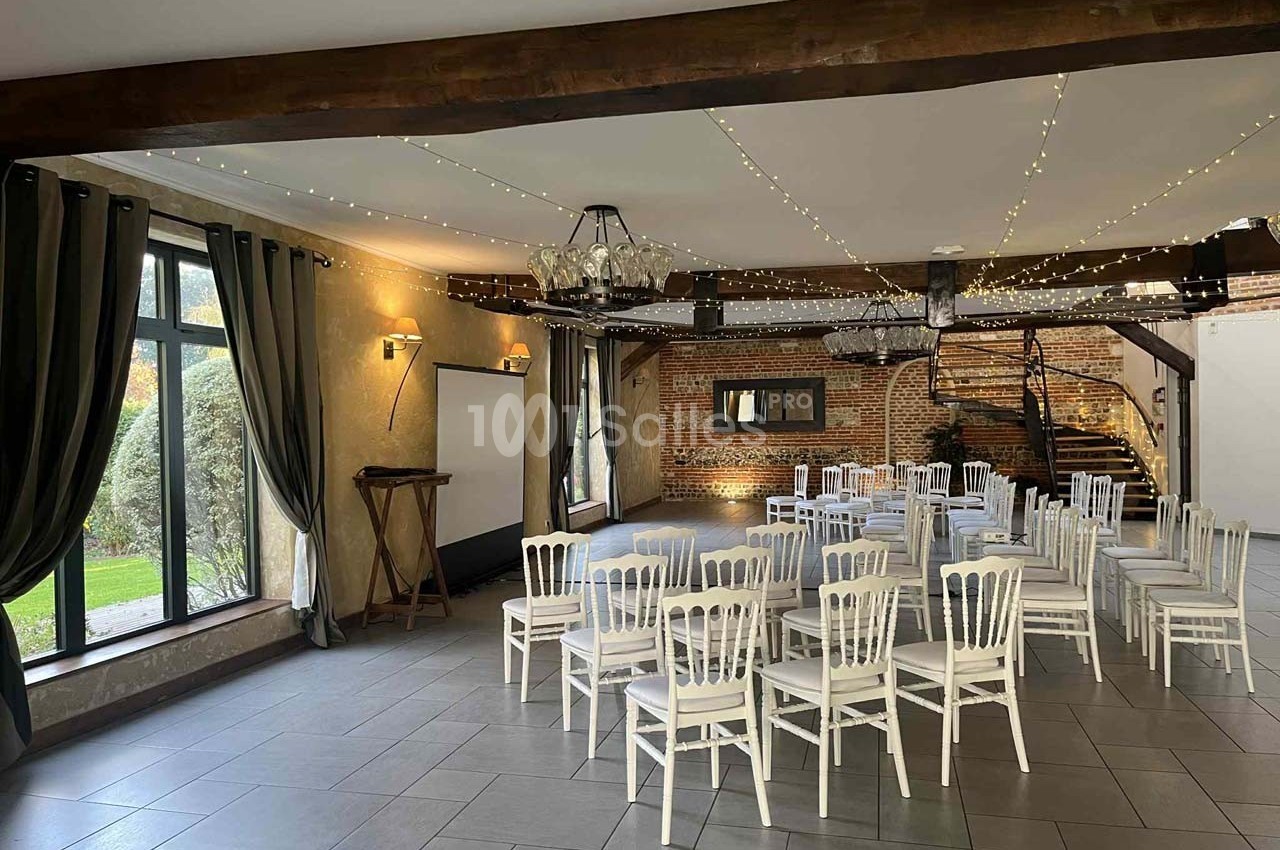 Salle lumineuse avec chaises blanches disposées en rangées, poutres apparentes, murs en briques et grandes fenêtres.