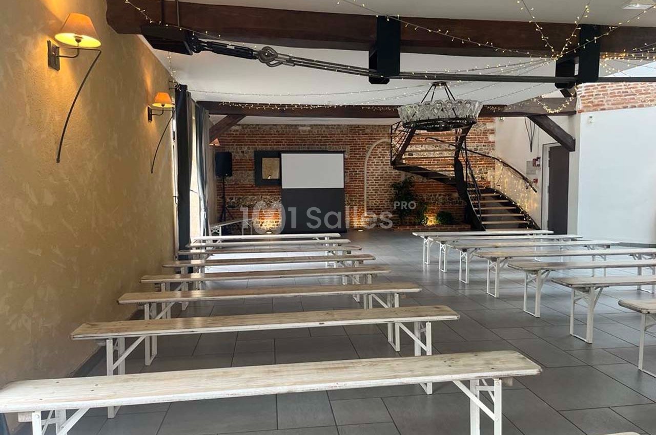 Salle lumineuse avec bancs en bois alignés, écran de projection, murs en briques et escalier en colimaçon.