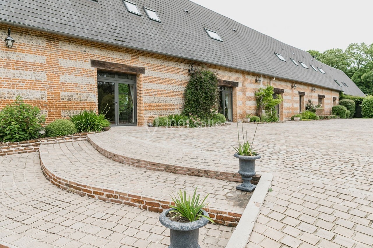 Façade en briques d'un bâtiment avec toit en ardoise, entourée d'une cour pavée et de plantes en pots.