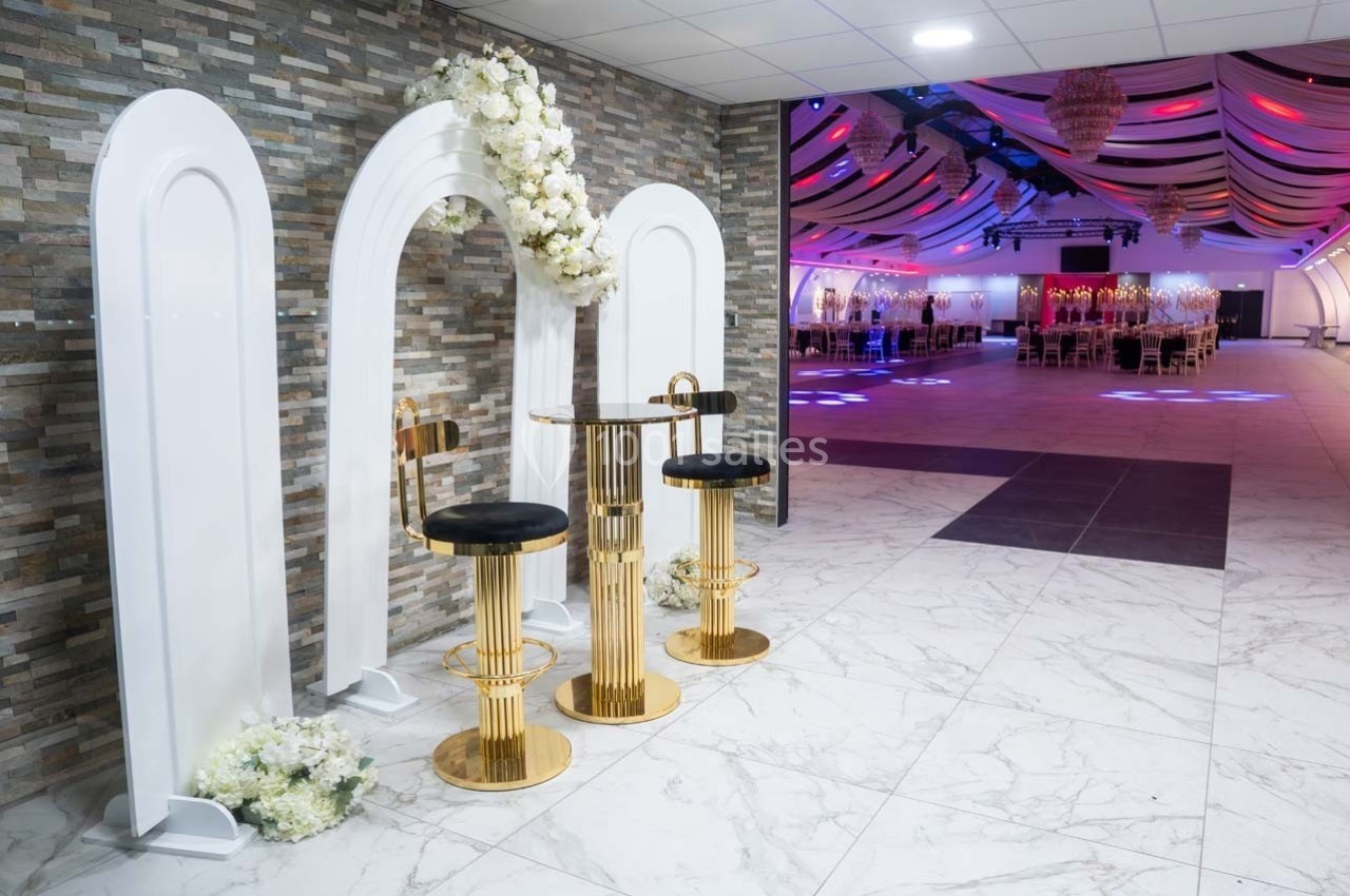 Deux tabourets dorés et une table assortie devant une arche décorée de fleurs blanches, dans une salle de réception.