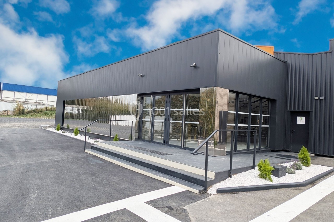 Façade d'un bâtiment moderne avec murs noirs, grandes baies vitrées et rampe d'accès, entouré d'un parking asphalté.