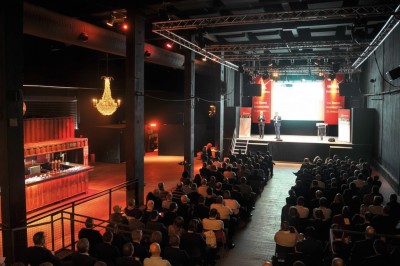 Foule dans une salle sombre éclairée par des lumières rouges intenses, avec une scène visible au fond.