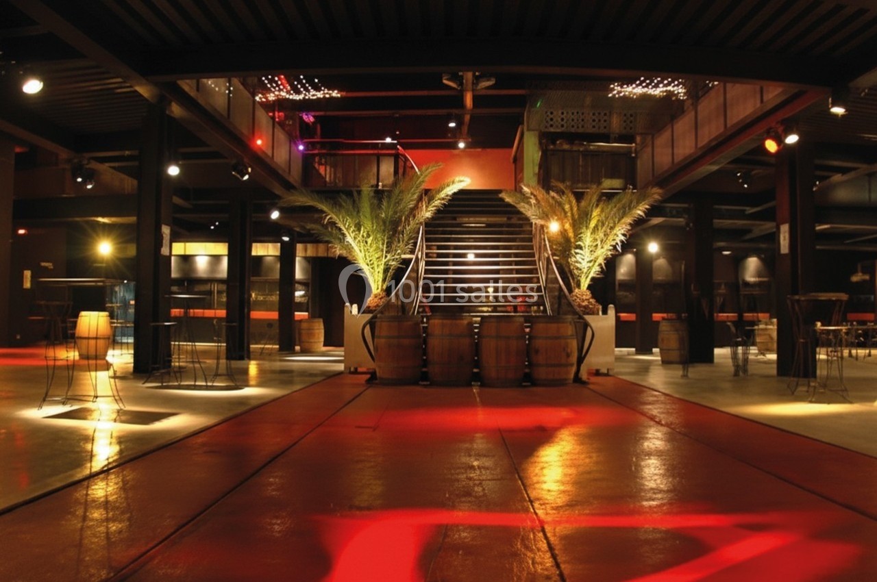 Salle spacieuse avec escalier central, éclairage tamisé, palmiers décoratifs et tonneaux en bois au premier plan.