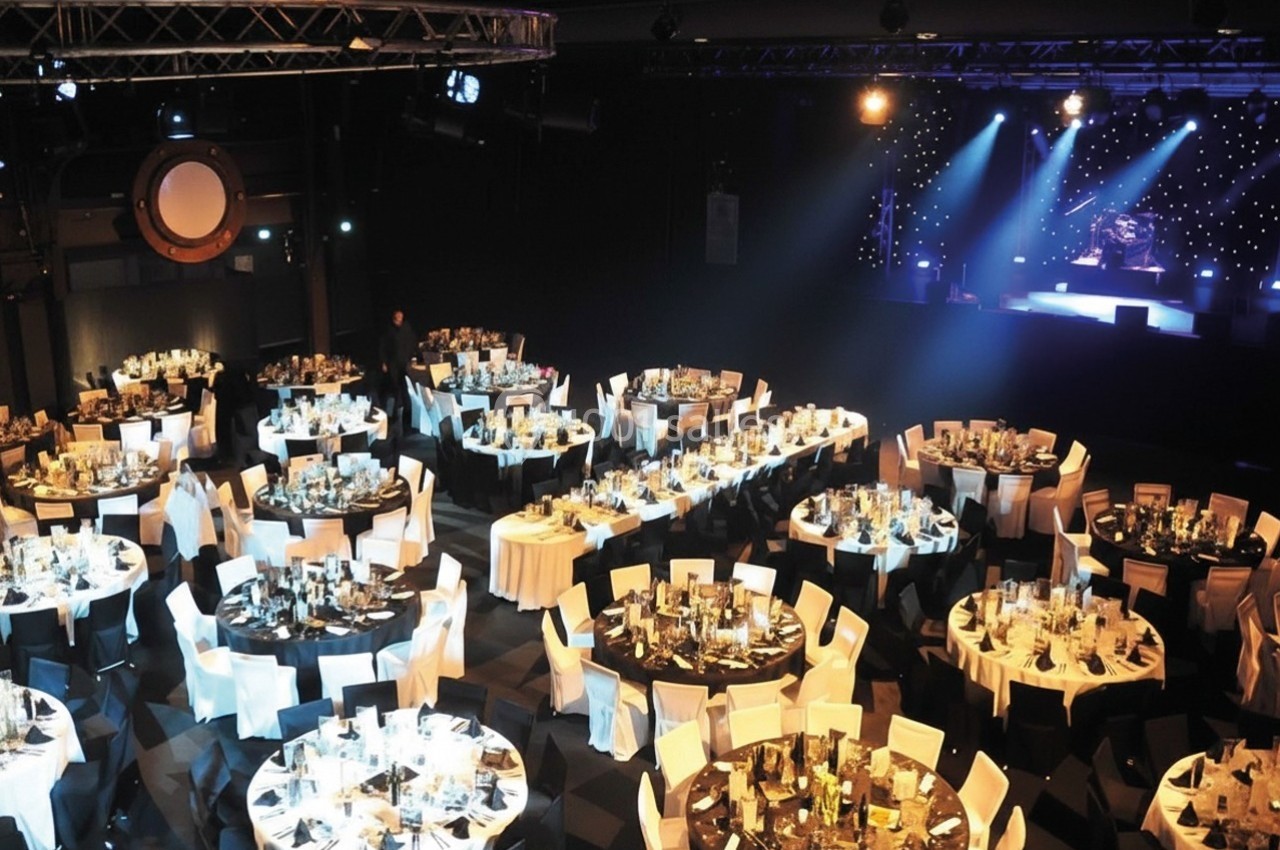 Salle de réception avec des tables rondes dressées pour un événement, éclairée par des projecteurs sur une scène.
