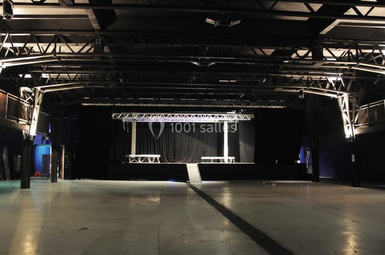 Salle de concert vide avec une scène surélevée, des structures métalliques au plafond et un éclairage tamisé.