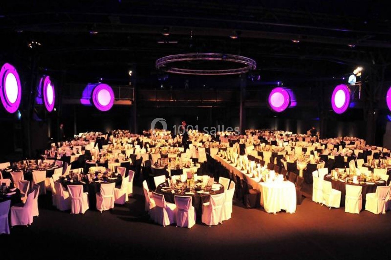 Salle de réception avec de nombreuses tables rondes dressées, éclairée par des lumières violettes suspendues.