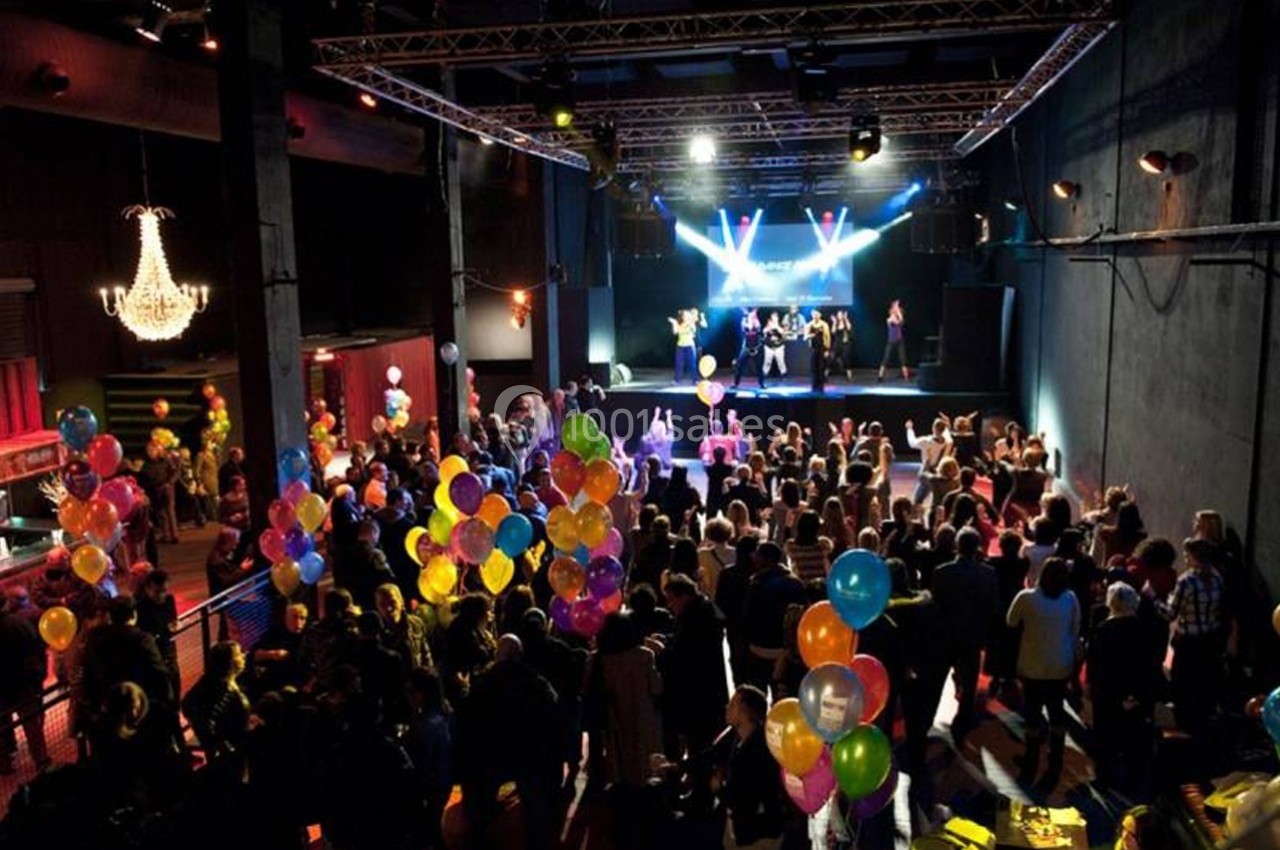 Salle de concert animée avec des spectateurs, des ballons colorés et une scène éclairée où des artistes se produisent.