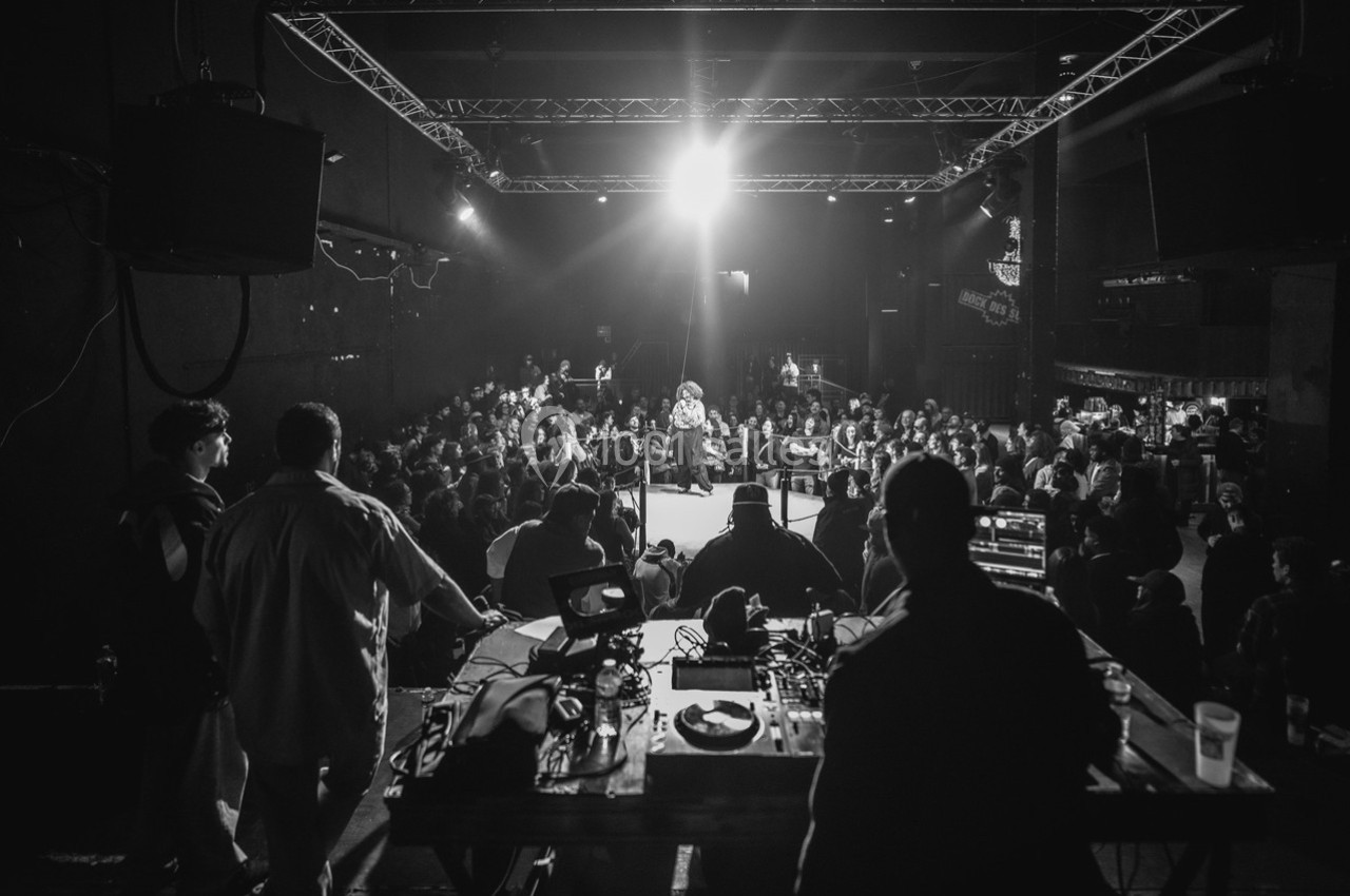 Vue en noir et blanc d'une salle de concert avec un artiste sur scène et un public attentif, éclairé par des projecteurs.