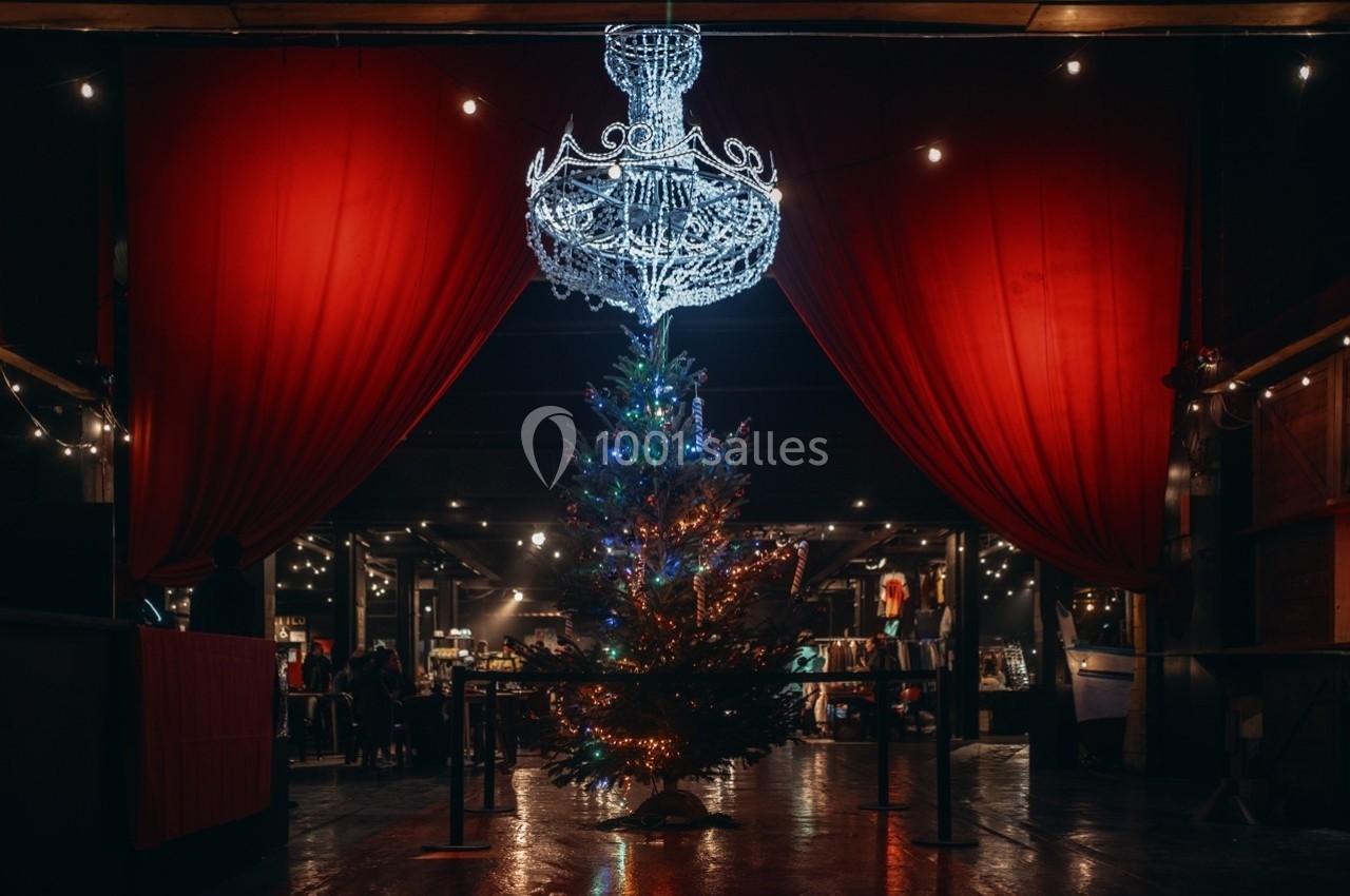 Sapin de Noël décoré sous un grand lustre illuminé, entouré de rideaux rouges dans une salle sombre.