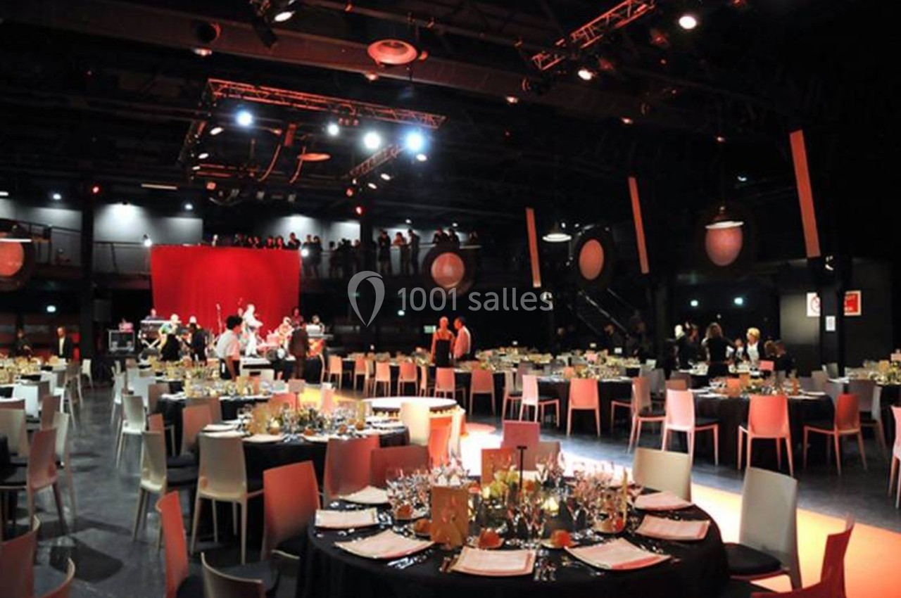 Salle de réception éclairée avec des tables dressées pour un événement, des invités et une scène en arrière-plan.