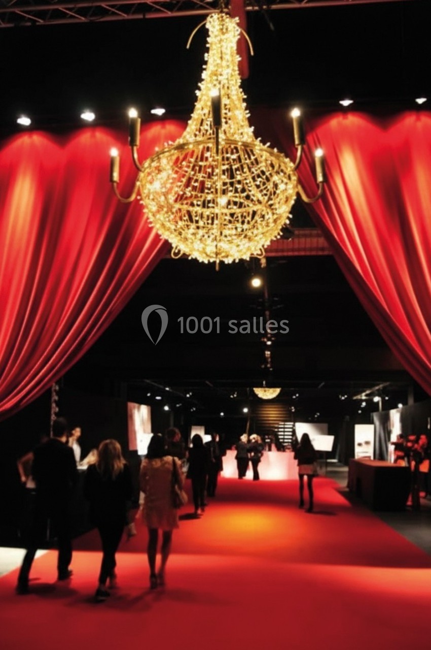 Un lustre doré suspendu au-dessus d'un tapis rouge entouré de rideaux rouges, avec des personnes marchant.