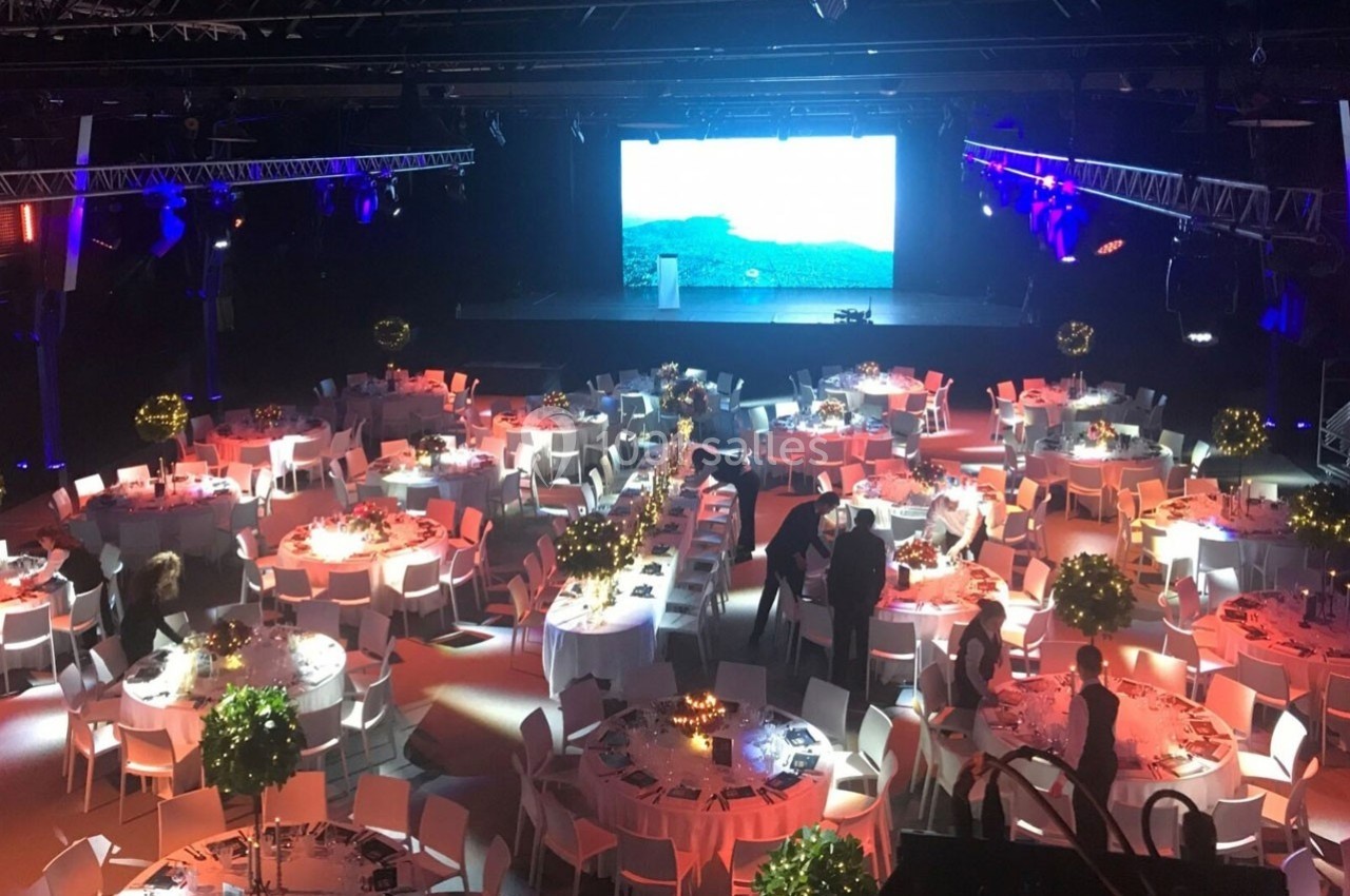 Salle de réception éclairée avec des tables rondes décorées, des invités et un écran géant en arrière-plan.