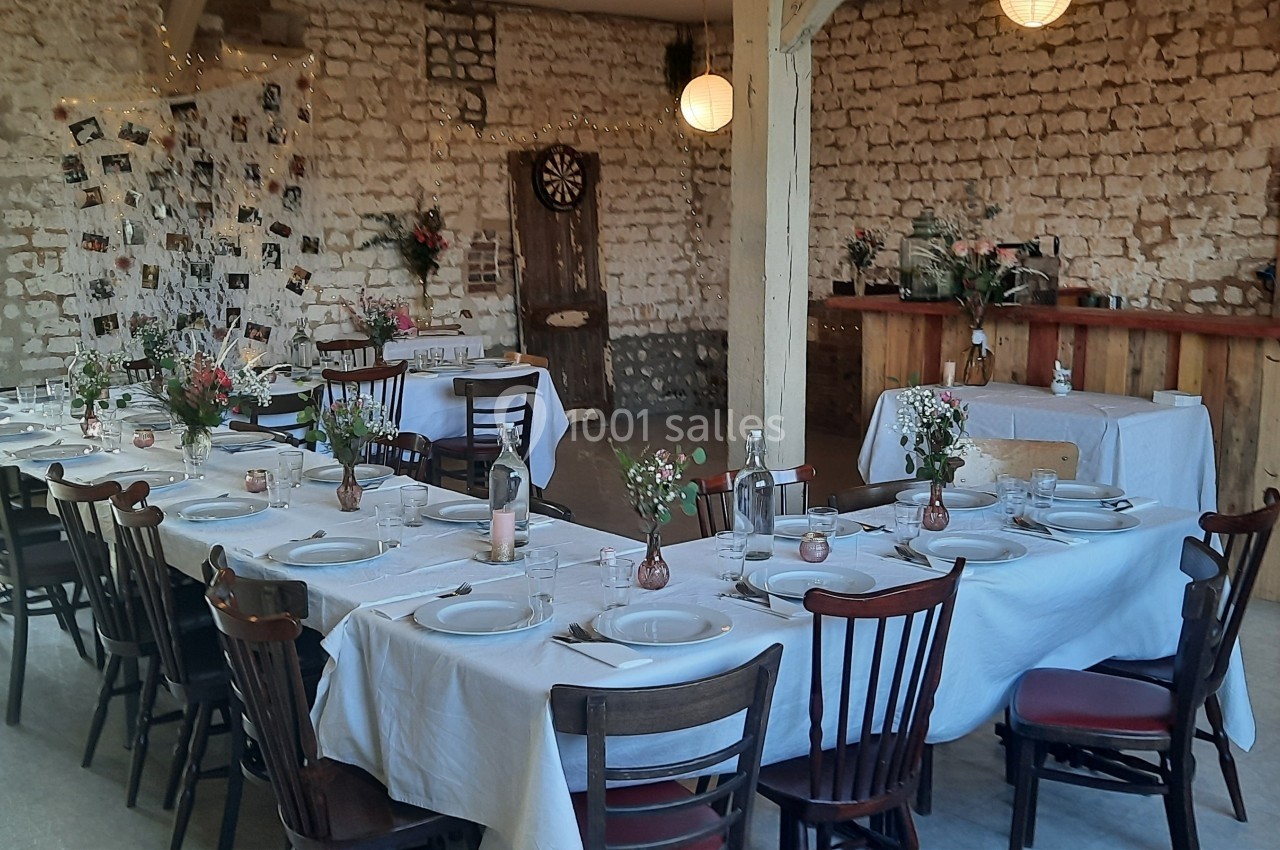 Salle rustique décorée pour un repas, avec tables dressées, nappes blanches, fleurs et murs en pierre apparente.