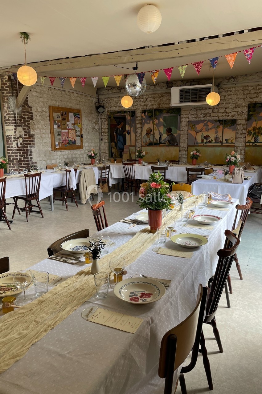 Salle décorée pour un repas festif avec longues tables dressées, guirlandes colorées et fleurs en centre de table.