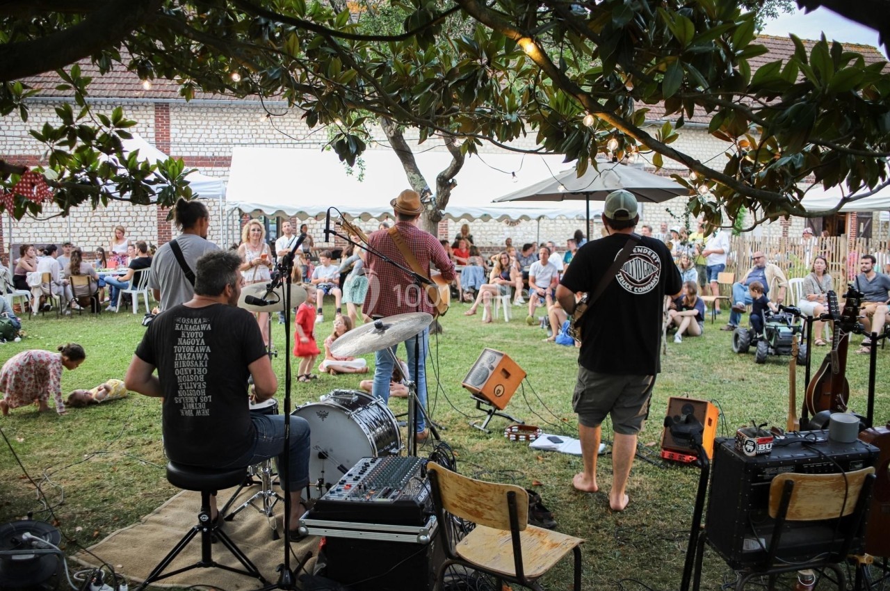 Groupe de musiciens jouant en plein air devant un public assis dans un jardin, entouré d'arbres et de bâtiments.