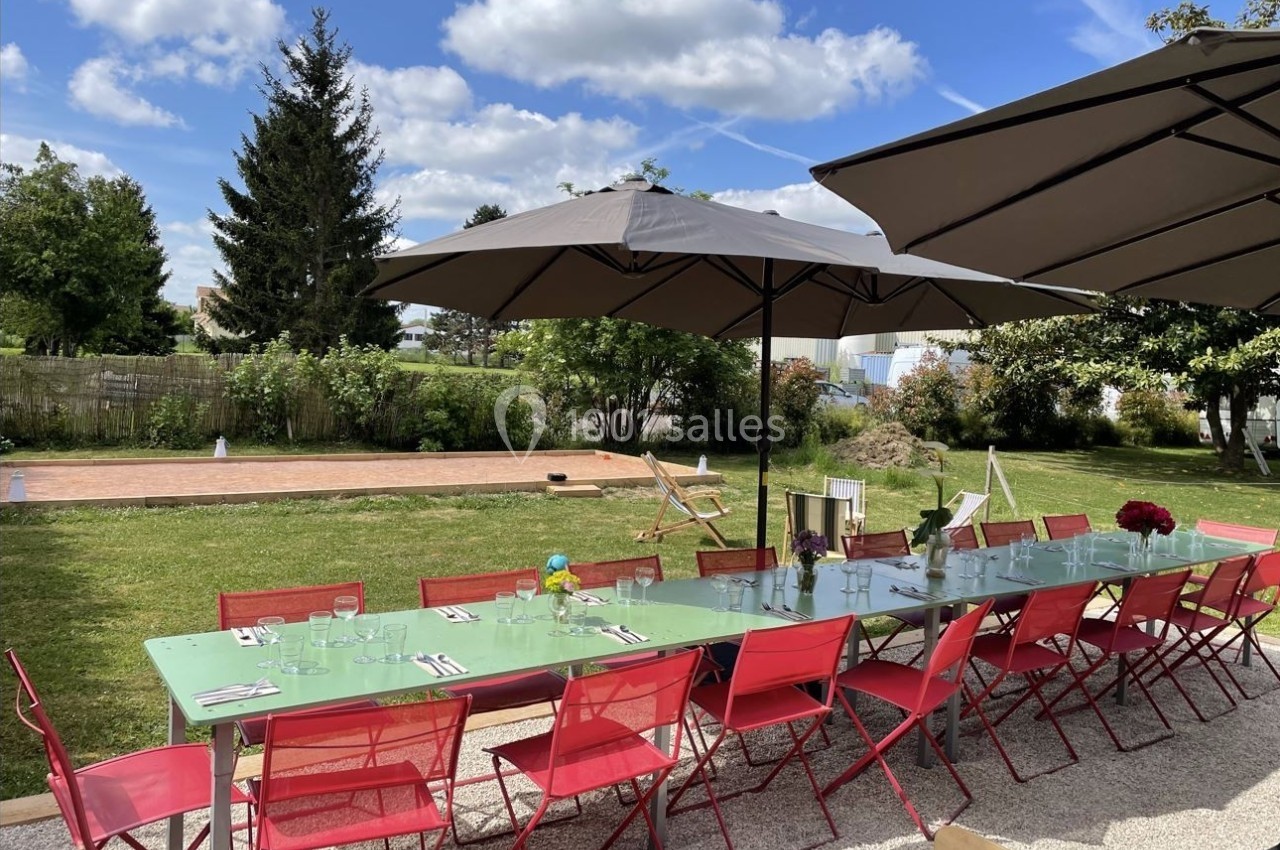 Table extérieure dressée avec chaises rouges et parasols, située dans un jardin sous un ciel partiellement nuageux.