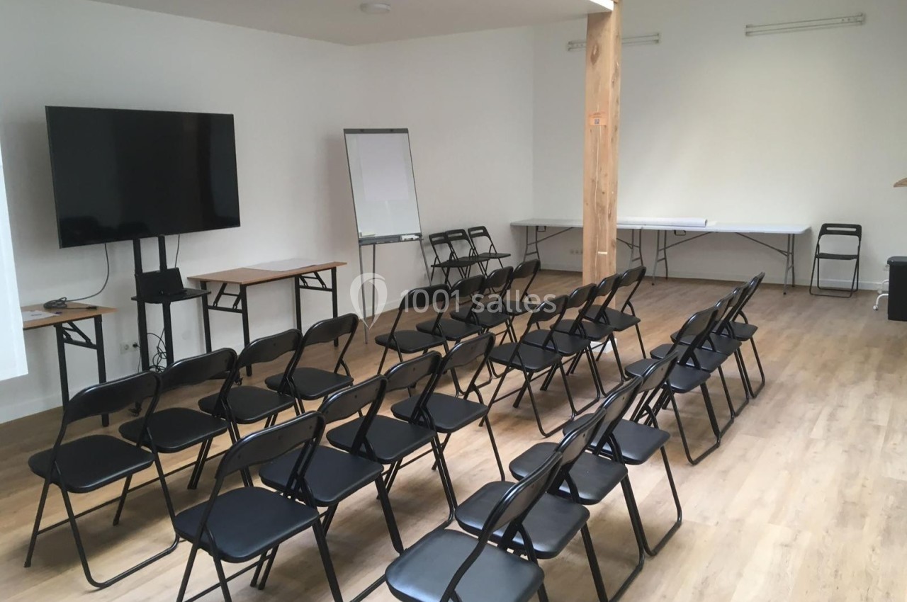 Salle de réunion avec chaises noires alignées, écran, tableau blanc et tables en fond sur un sol en bois clair.