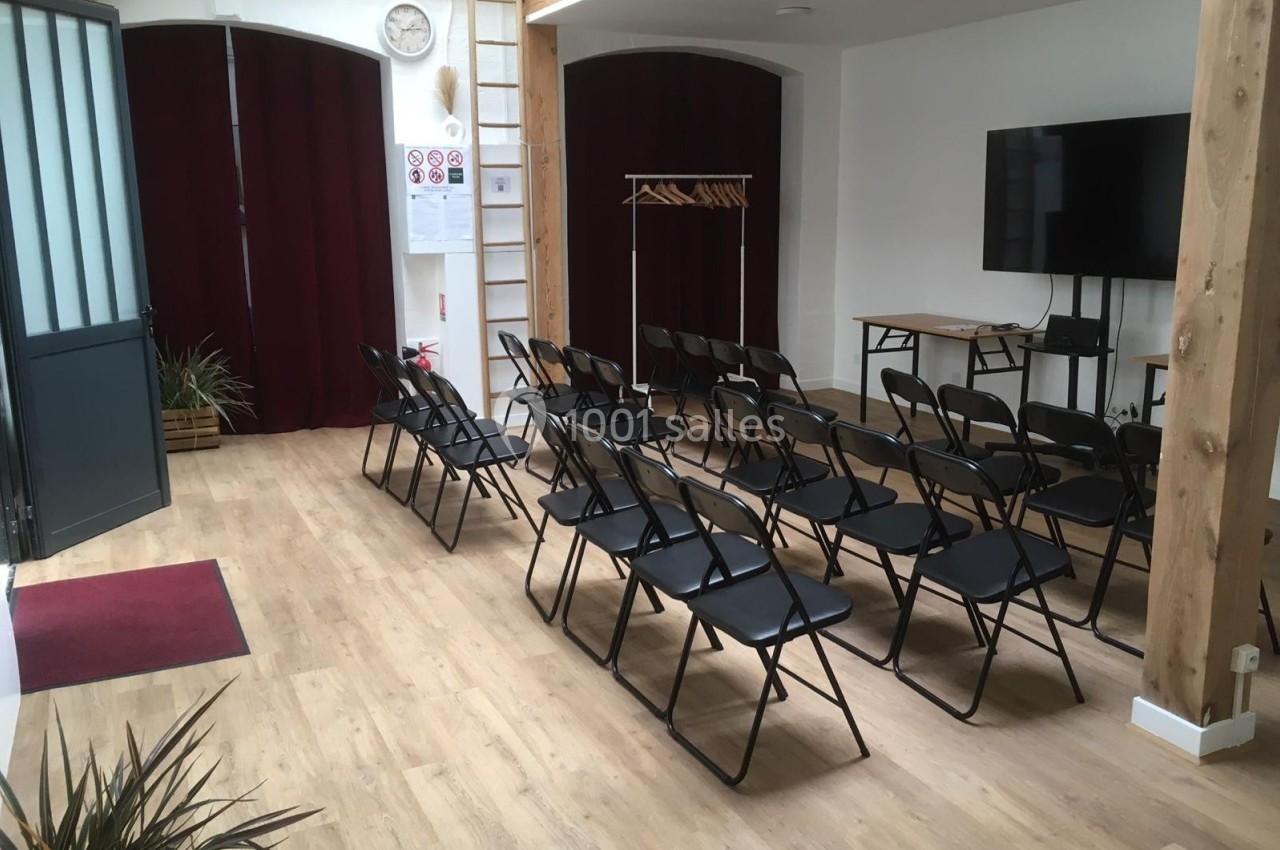 Salle lumineuse avec chaises pliantes alignées, écran, porte ouverte et rideaux bordeaux en arrière-plan.