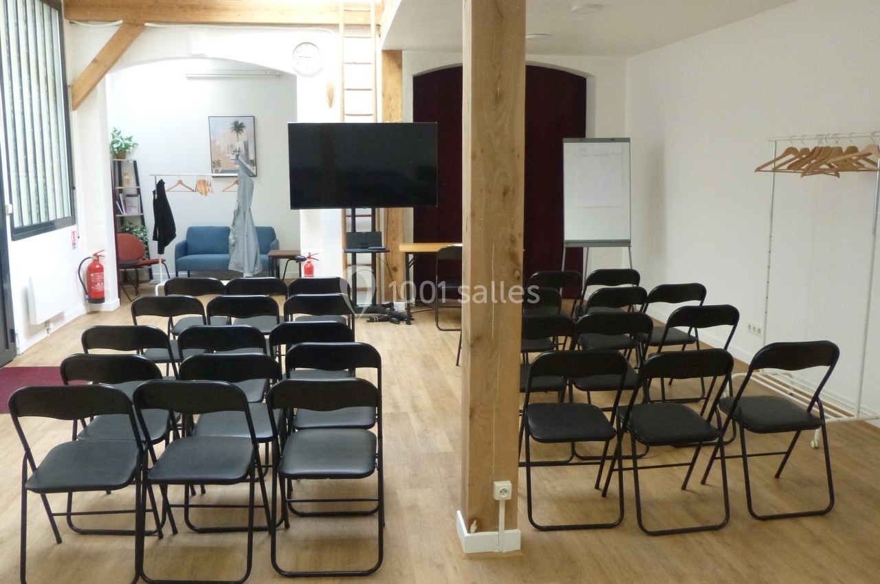 Salle de réunion avec chaises noires alignées, écran, paperboard, portants et mobilier dans un espace lumineux.